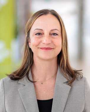 Eine Frau mit langen, braunen Haaren trägt einen grauen Blazer und ein schwarzes Oberteil. Sie lächelt leicht und steht vor einem unscharfen Hintergrund mit hellen Farben.